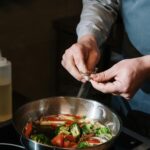 A chef seasons vegetables in a frying pan on a modern stovetop indoors.