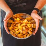 From above of crop unrecognizable person in apron holding plate with pieces of delicious fried potato